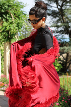 A woman wearing a vibrant red feather-trimmed shawl over a black top, photographed outdoors in a garden setting with trees and a building in the background.