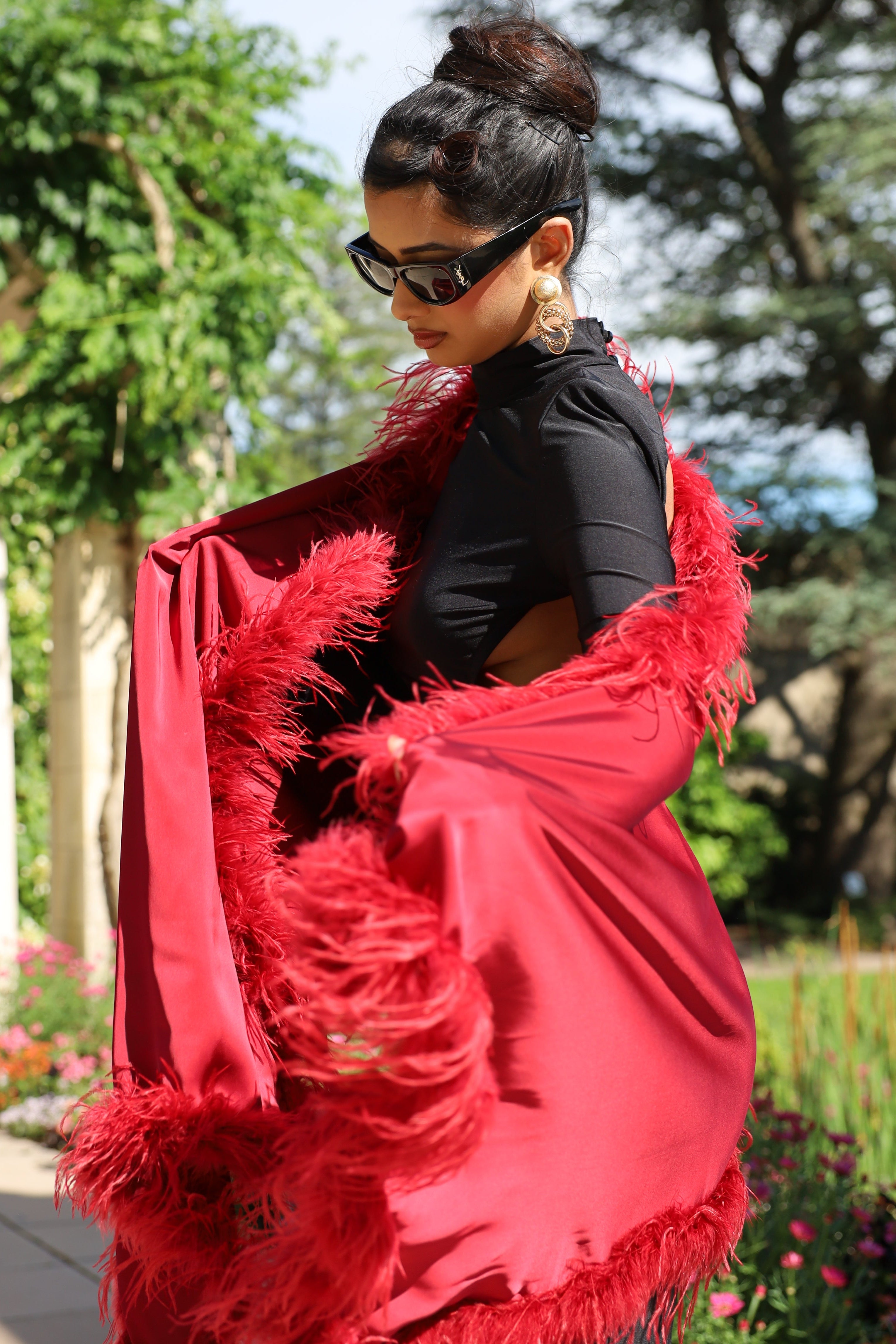 A woman wearing a vibrant red feather-trimmed shawl over a black top, photographed outdoors in a garden setting with trees and a building in the background.